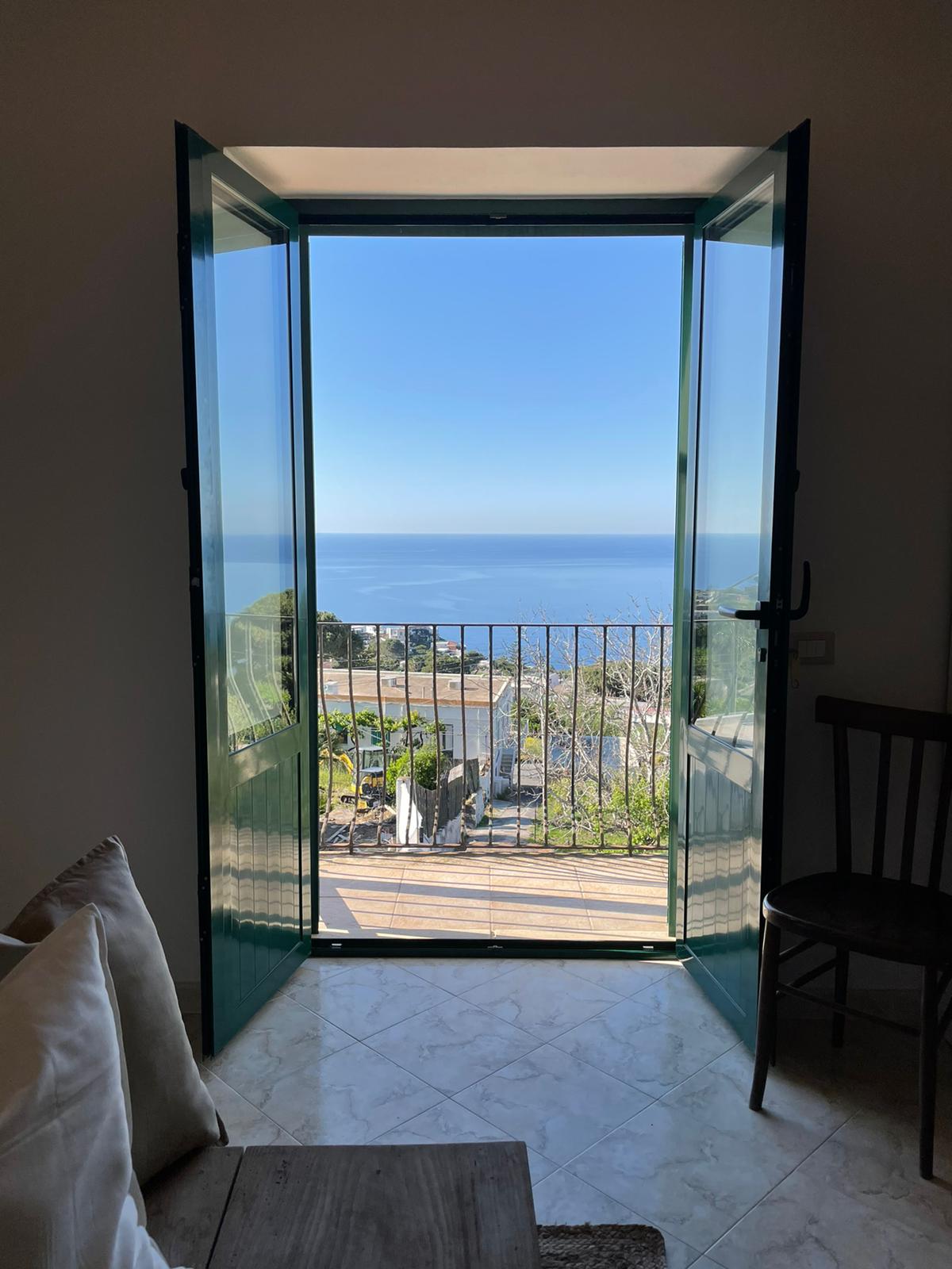 The image features an open window with green shutters, providing an unobstructed view of the serene blue sea and coastal landscape. A small balcony with a railing enhances the scenic outlook, while a glimpse of the interior living space is visible with light-colored furnishings.