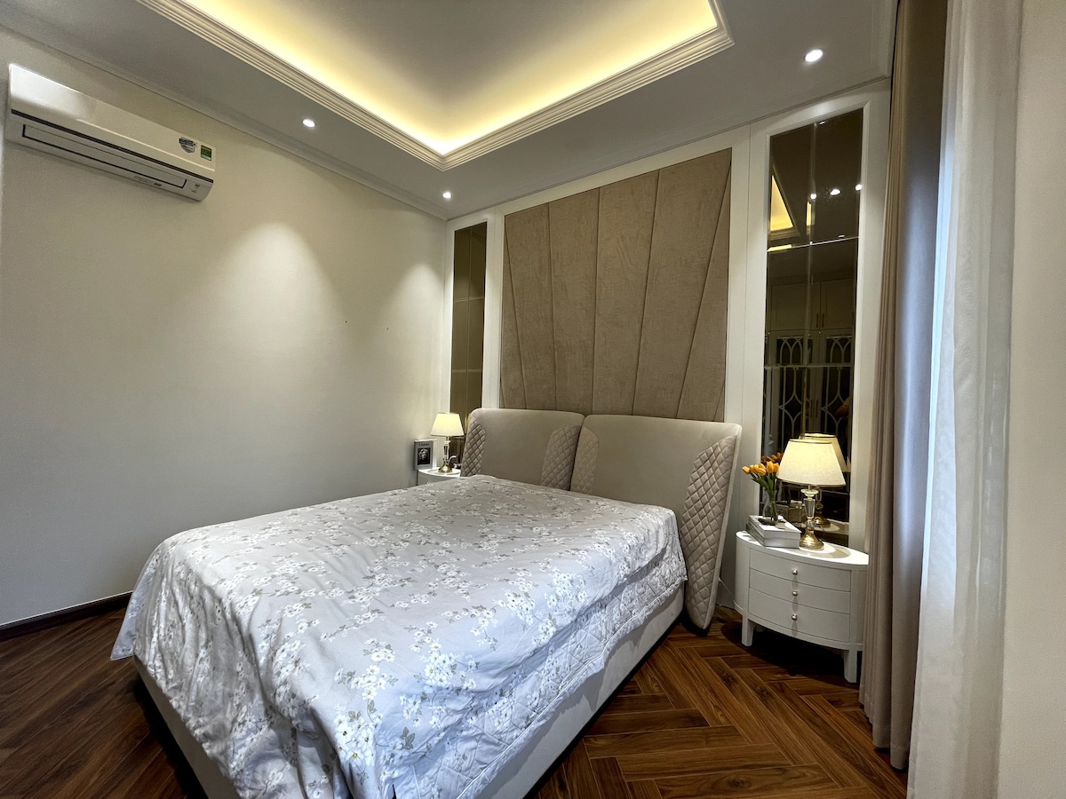 A spacious bedroom features a plush headboard framing a large bed covered in a soft, light-colored comforter. Nightstands with lamps are placed on either side of the bed. An air conditioning unit is mounted on the wall, and natural light enters through sheer curtains.