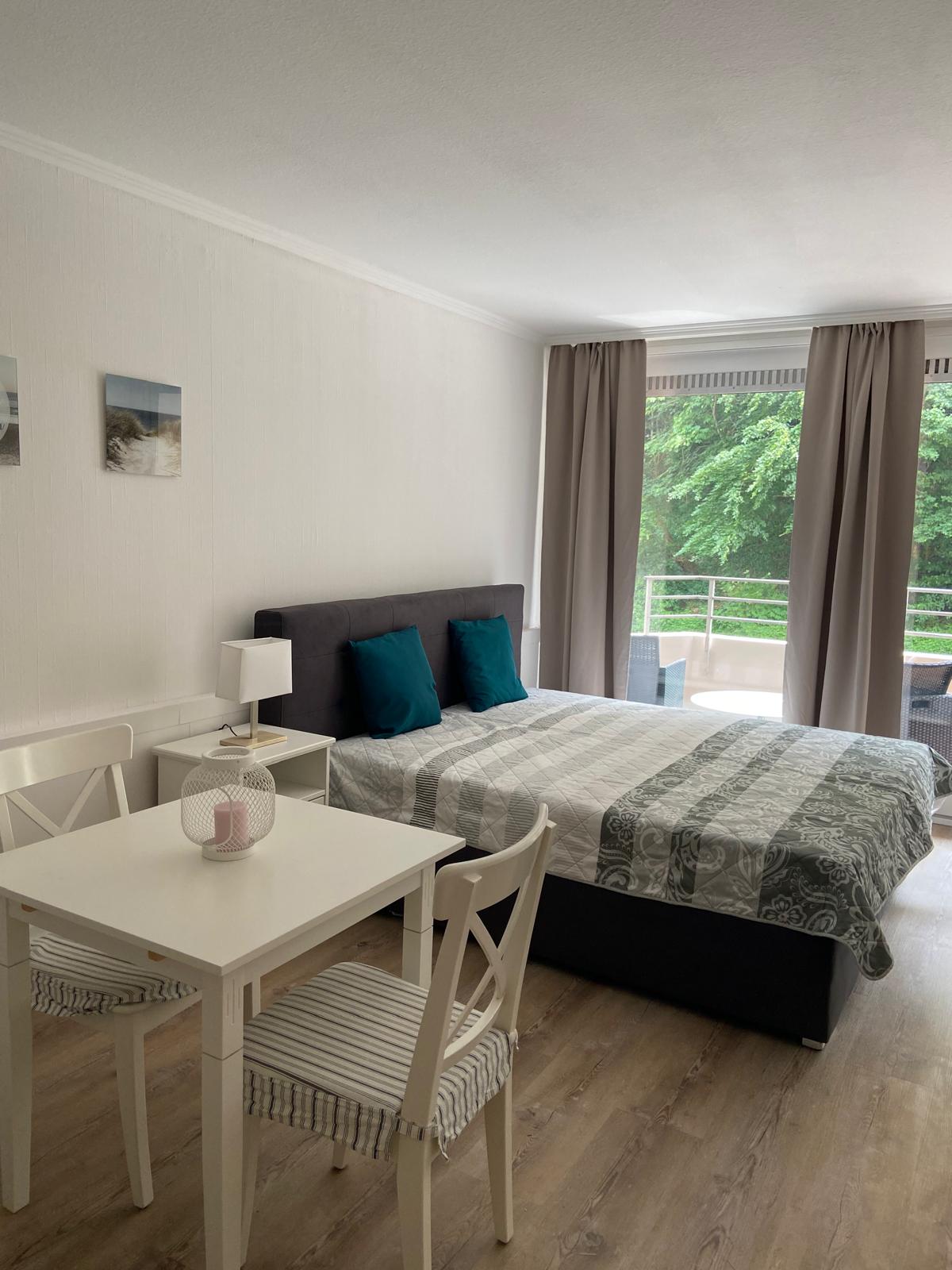 A bright and airy living space features a large double bed with a patterned duvet and teal accent pillows. A small dining table is set next to the bed, accompanied by two chairs. Sunlight streams through large windows adorned with light drapes, revealing green outdoor foliage.