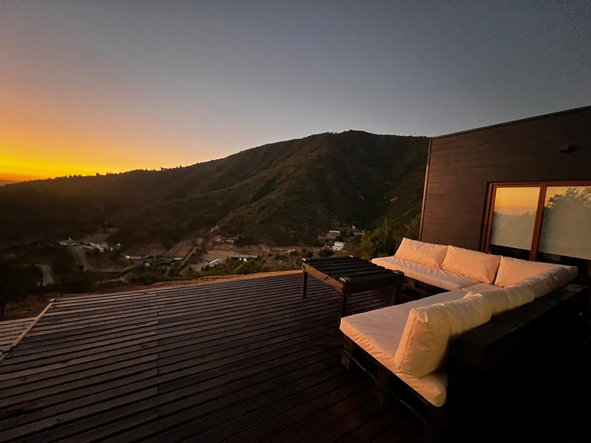 A spacious outdoor seating area is set on a wooden deck, offering comfortable cushions for relaxation. The view showcases rolling hills bathed in sunset colors, with the horizon glowing in shades of orange and purple. Large windows reflect the serene landscape.