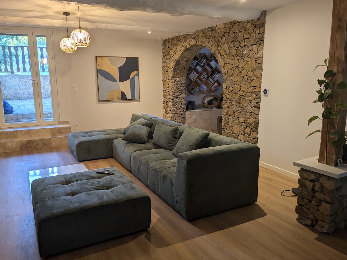 A modern living area features a spacious sectional sofa in soft green hues, complemented by a matching ottoman. Natural light enters through large sliding doors, highlighting a stone archway that adds character to the room. Minimal decor is present, creating a calming environment.