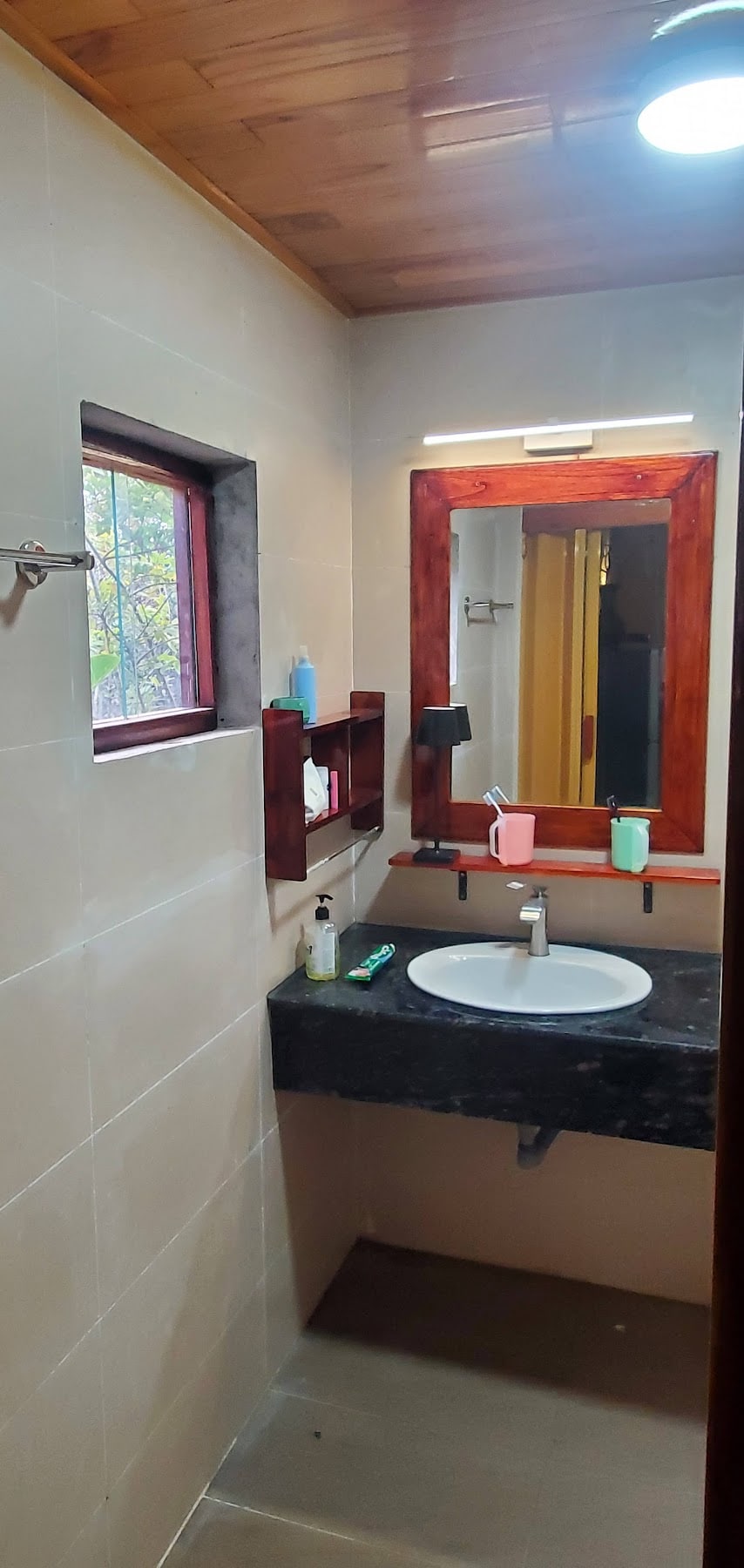 A well-appointed bathroom features a round sink atop a dark stone countertop. A wooden mirror frame complements the decor. Shelves hold toiletries, while natural light filters through a small window, illuminating the space.