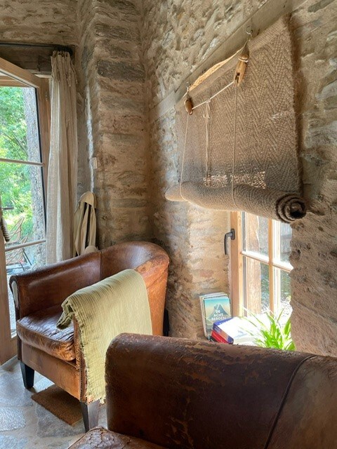 A cozy corner showcases two brown leather chairs positioned near a window, with natural light filtering through. A woven shade covers the window, and a small potted plant adds a touch of greenery. A soft blanket drapes over one chair, enhancing the inviting atmosphere.