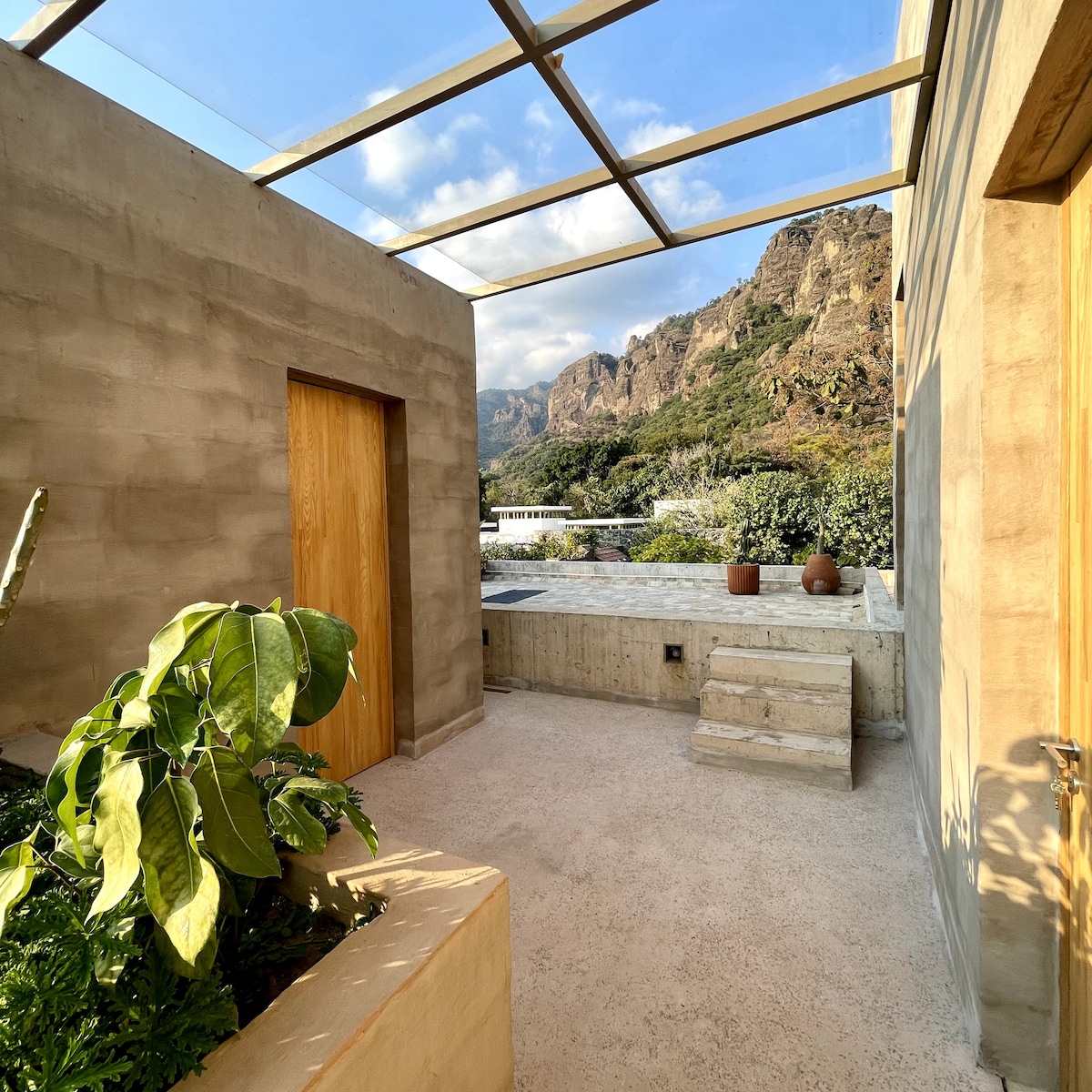 An outdoor terrace features a mix of natural stone and concrete surfaces, complemented by greenery. Large glass panels provide views of the surrounding mountains, enhancing the connection to nature. A wooden door and clay pots are also visible, indicating a blend of materials.
