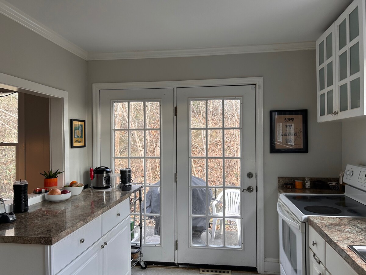 A bright kitchen area features white cabinets and a granite countertop. Double glass doors allow natural light to enter, revealing a view of the outside. Various kitchen appliances, including a coffee maker and stove, are neatly arranged within the space.