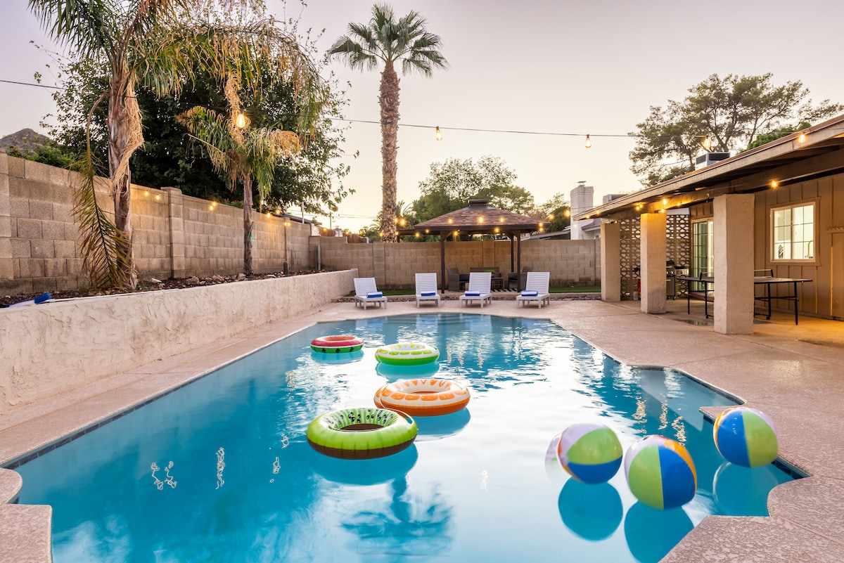 A private pool shines under the evening sky, surrounded by palm trees and lit by string lights. Colorful pool floats and beach balls add a playful touch to the water. Lounge chairs provide inviting seating along the poolside, enhancing the outdoor retreat experience.