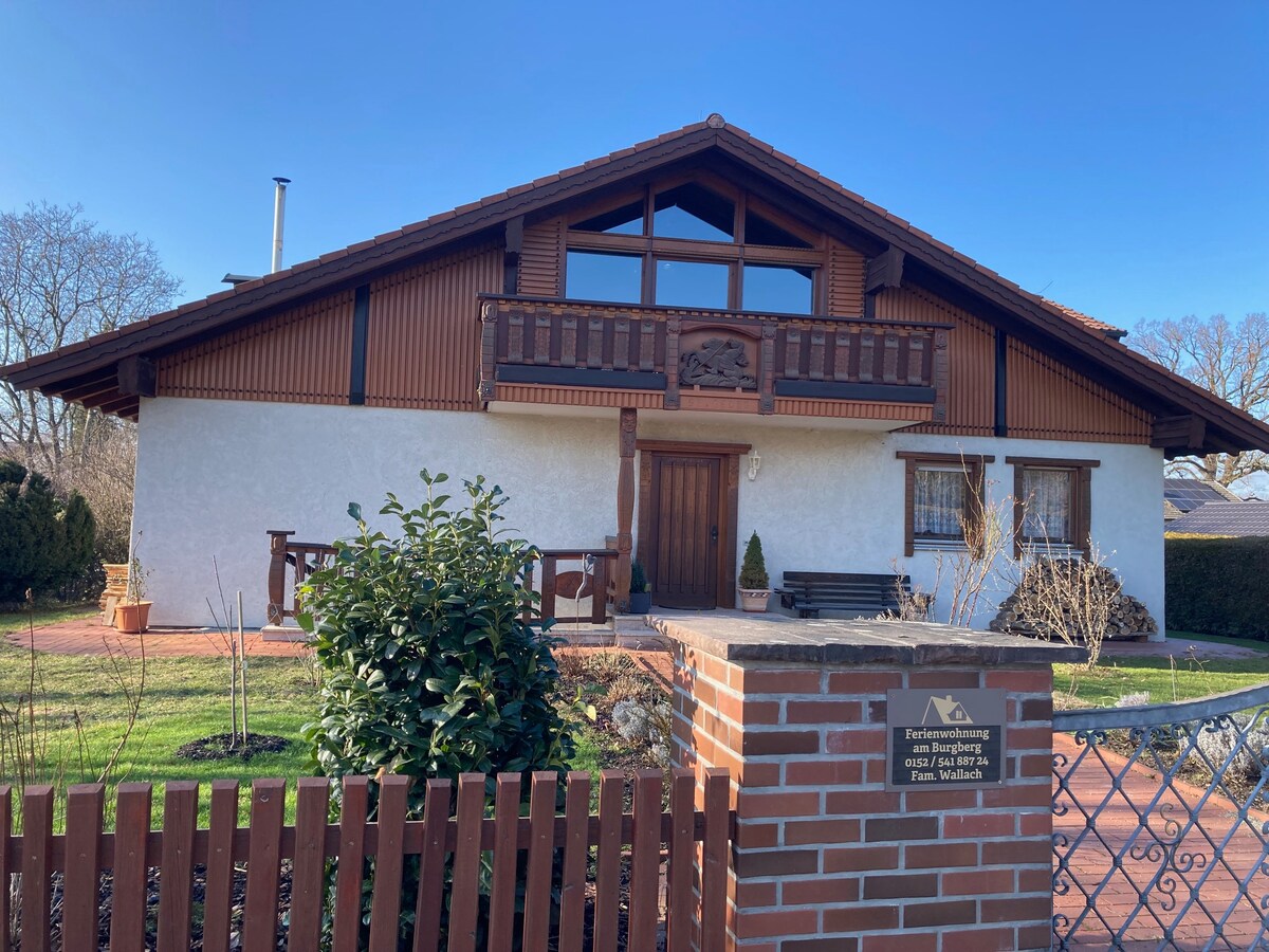 A traditional chalet-style building features a sloped roof with wooden balcony accents. The exterior is a mix of white stucco and natural wood, surrounded by manicured gardens and a stone pathway. A welcoming entrance is framed by a low brick fence and a wrought iron gate.