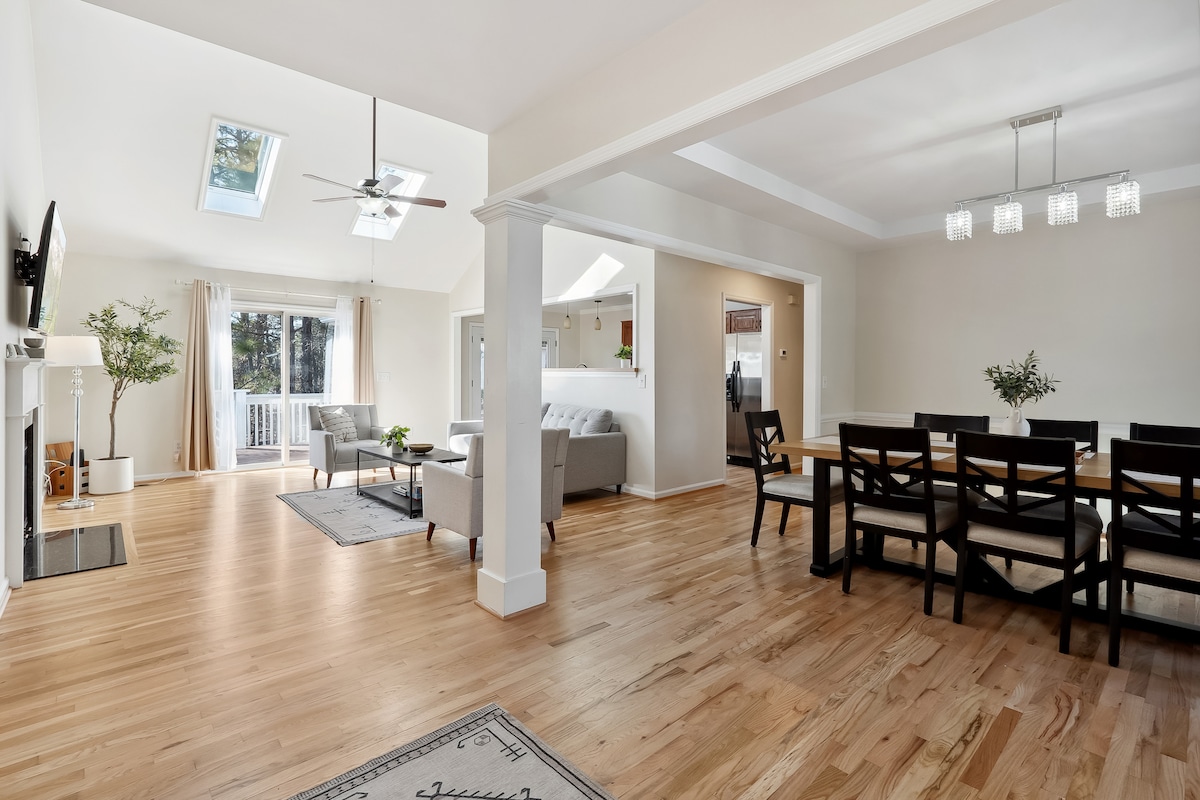 An open-concept living space features hardwood flooring and large windows allowing natural light to fill the room. A comfortable seating area is located near the fireplace, while a dining area with a large table accommodates several guests. Skylights add a bright touch to the room.