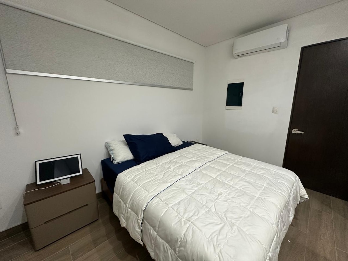 The sleeping area features a spacious bed with a light-colored quilt, accented by dark blue and white pillows. A bedside table is positioned next to the bed, and a small electronic device sits atop it. A wall-mounted air conditioning unit is visible, contributing to the room's comfort.