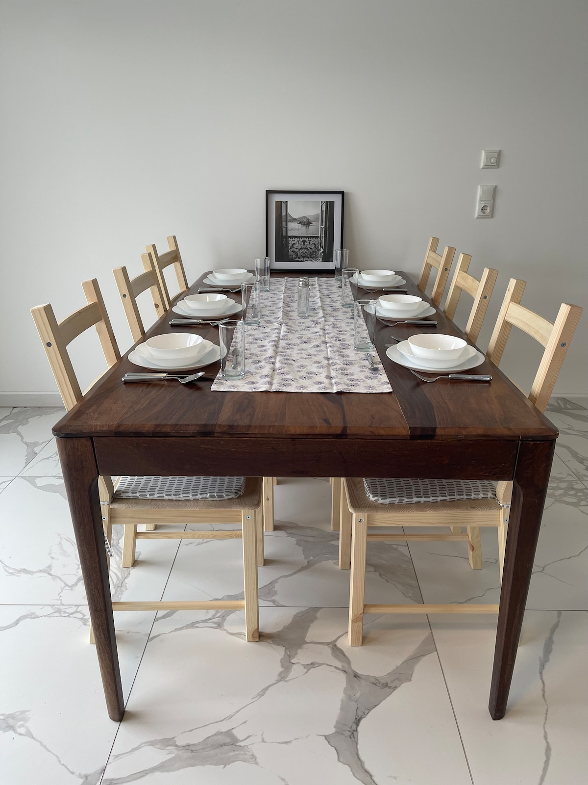 A long wooden dining table is set for eight with white plates, silverware, and glasses arranged neatly. A light-colored table runner decorates the center, and a framed black-and-white photograph adorns the wall behind the table. Simple wooden chairs with wicker seats complement the setting.