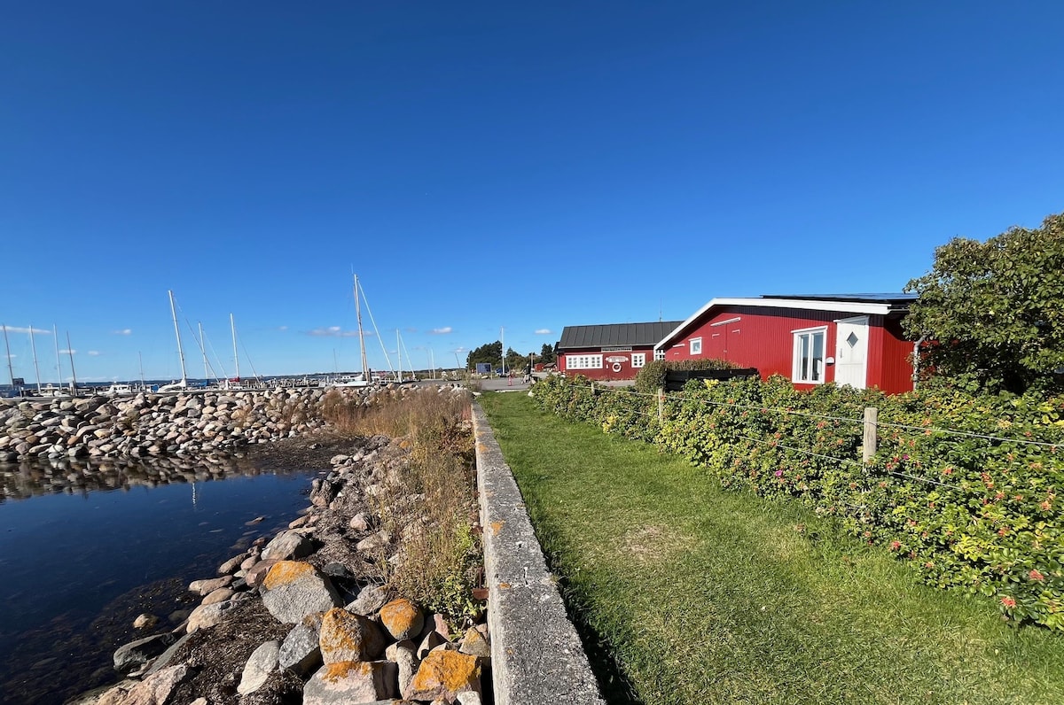 A red house sits alongside a waterfront path surrounded by a lush green lawn. The shoreline features stones and a calm body of water, with several sailboats visible in the distance under a clear blue sky.
