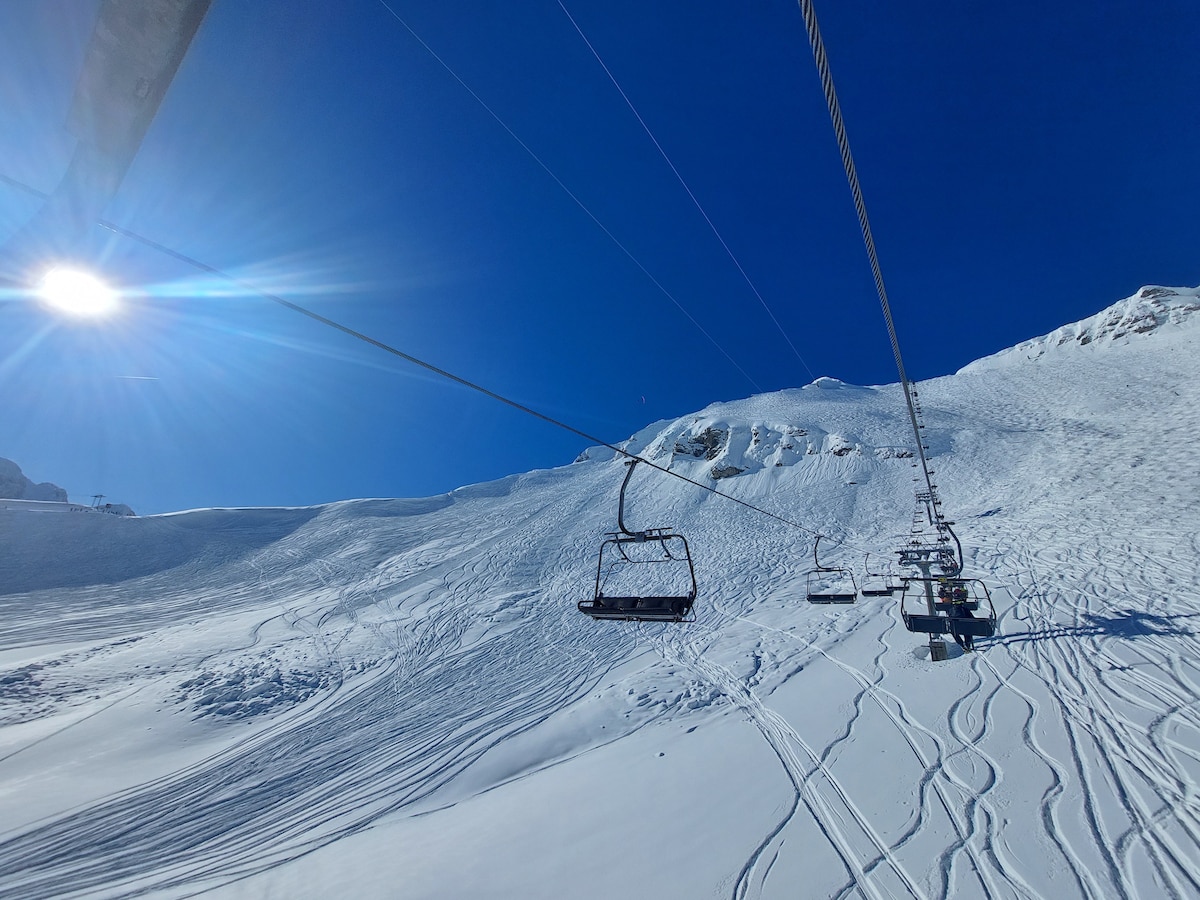 A ski lift ascends over a snow-covered slope, surrounded by a clear blue sky. The area shows tracks suggesting recent activity, while the sun reflects brightly off the snow, creating a serene winter atmosphere.