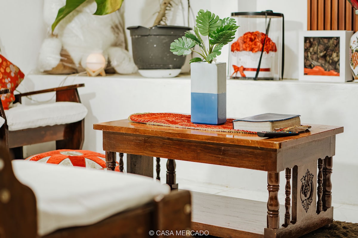 A wooden coffee table is positioned at the center of the room, adorned with a colorful runner and a ceramic vase holding greenery. Two upholstered chairs with light cushions are visible, alongside decorative accents in the background, creating a cozy and inviting space.