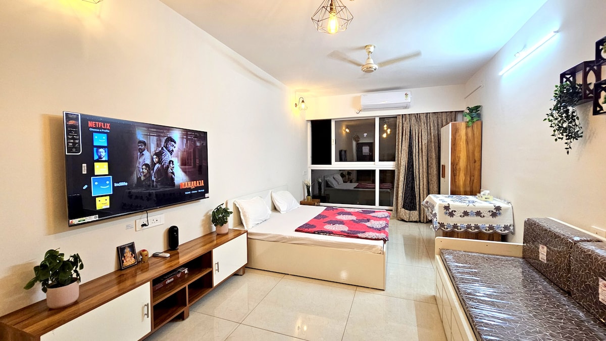 A spacious studio apartment features an ultra king-size bed, a sofa bed, and a large wall-mounted TV displaying streaming content. Soft natural light enters through a window, creating a bright atmosphere. A cozy dining area with a table is visible near the window.