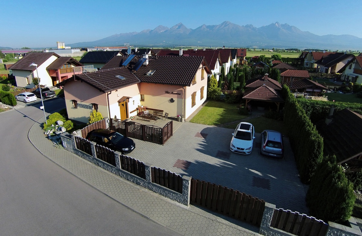 An aerial view shows a residential area with various houses and well-maintained gardens. The foreground features a spacious parking area with multiple vehicles. The backdrop includes the towering High Tatras mountains, providing a scenic landscape.
