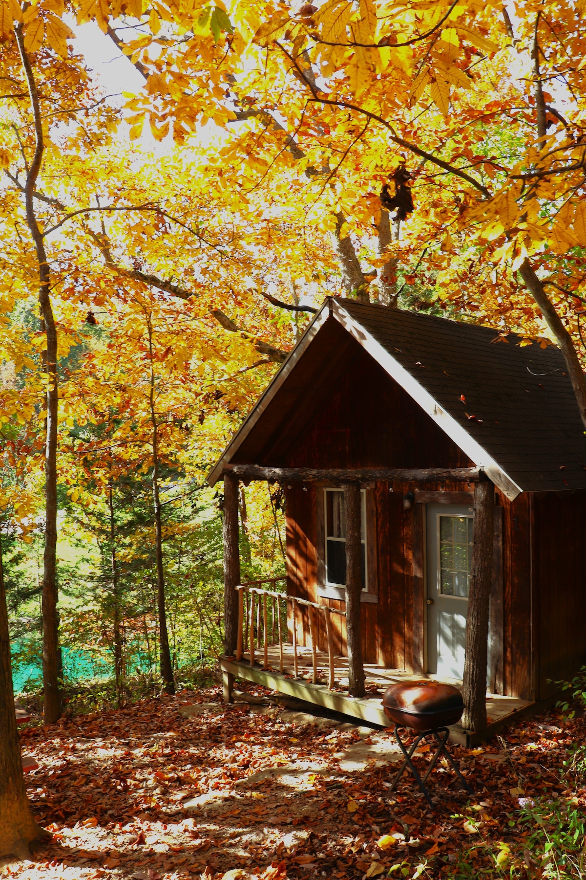 A rustic cabin is nestled among autumn-colored trees, featuring a small porch with wooden railings. Leaves cover the ground, creating a natural setting. A grill is positioned on the porch, and sunlight filters through the vibrant foliage, providing a warm atmosphere.
