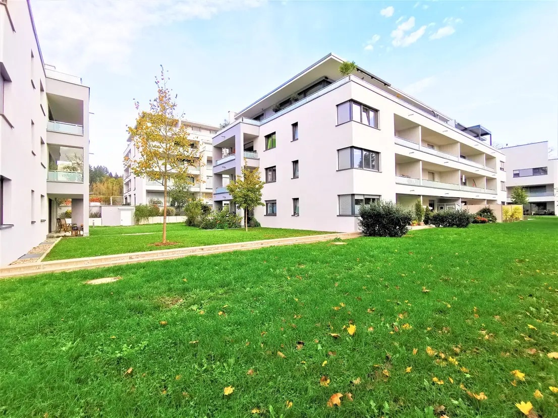 A modern multi-story building is surrounded by a well-kept green lawn, featuring clusters of shrubs and trees. Multiple balconies are visible on the upper floors, while large windows reflect natural light. The serene environment suggests a peaceful retreat in the landscape.