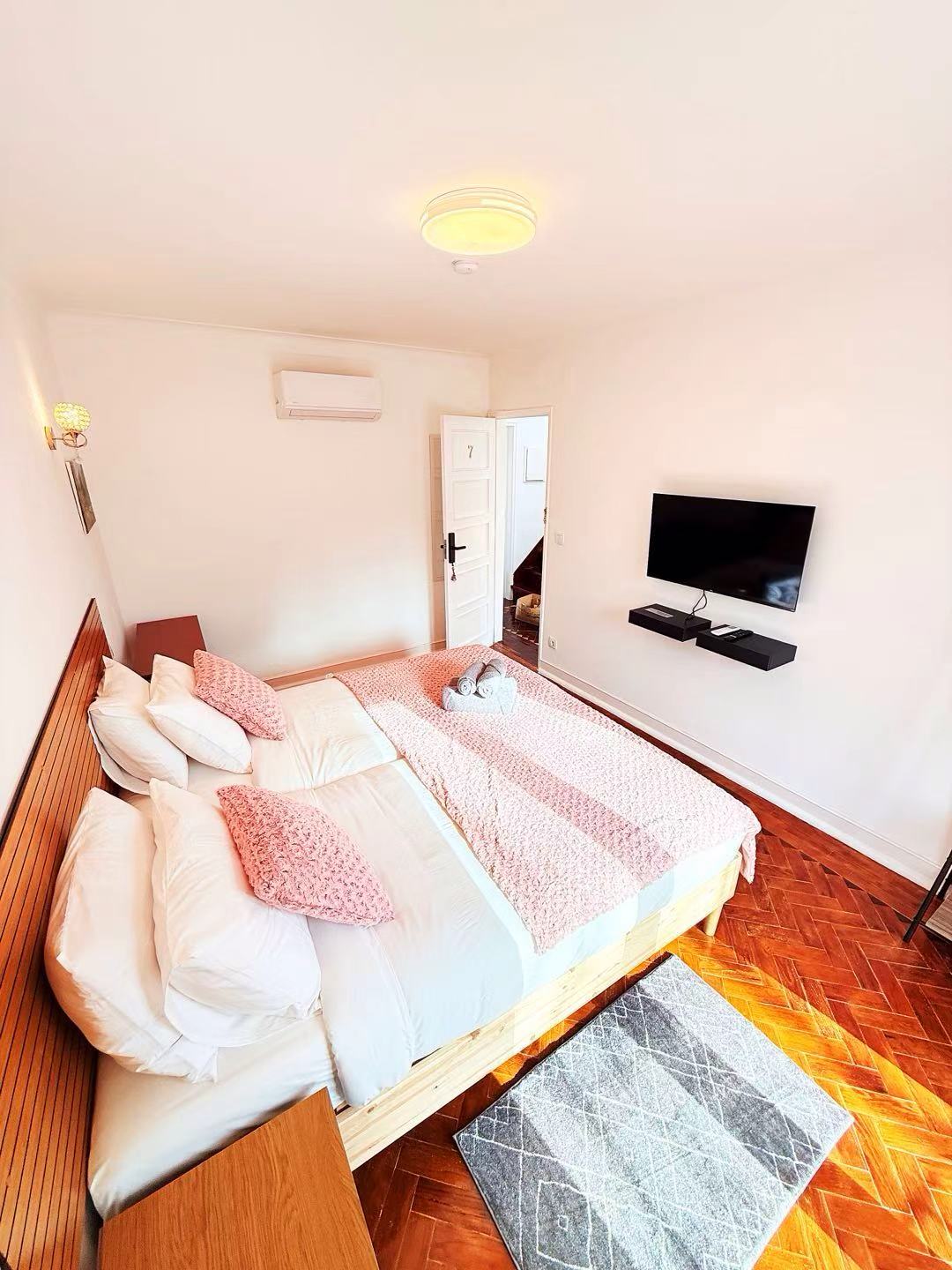 A well-lit bedroom is showcased with a spacious bed dressed in soft linens and decorative pillows. A Smart TV is mounted on the wall opposite the bed. Natural light streams through a window, illuminating the wooden parquet flooring and minimal furnishings.
