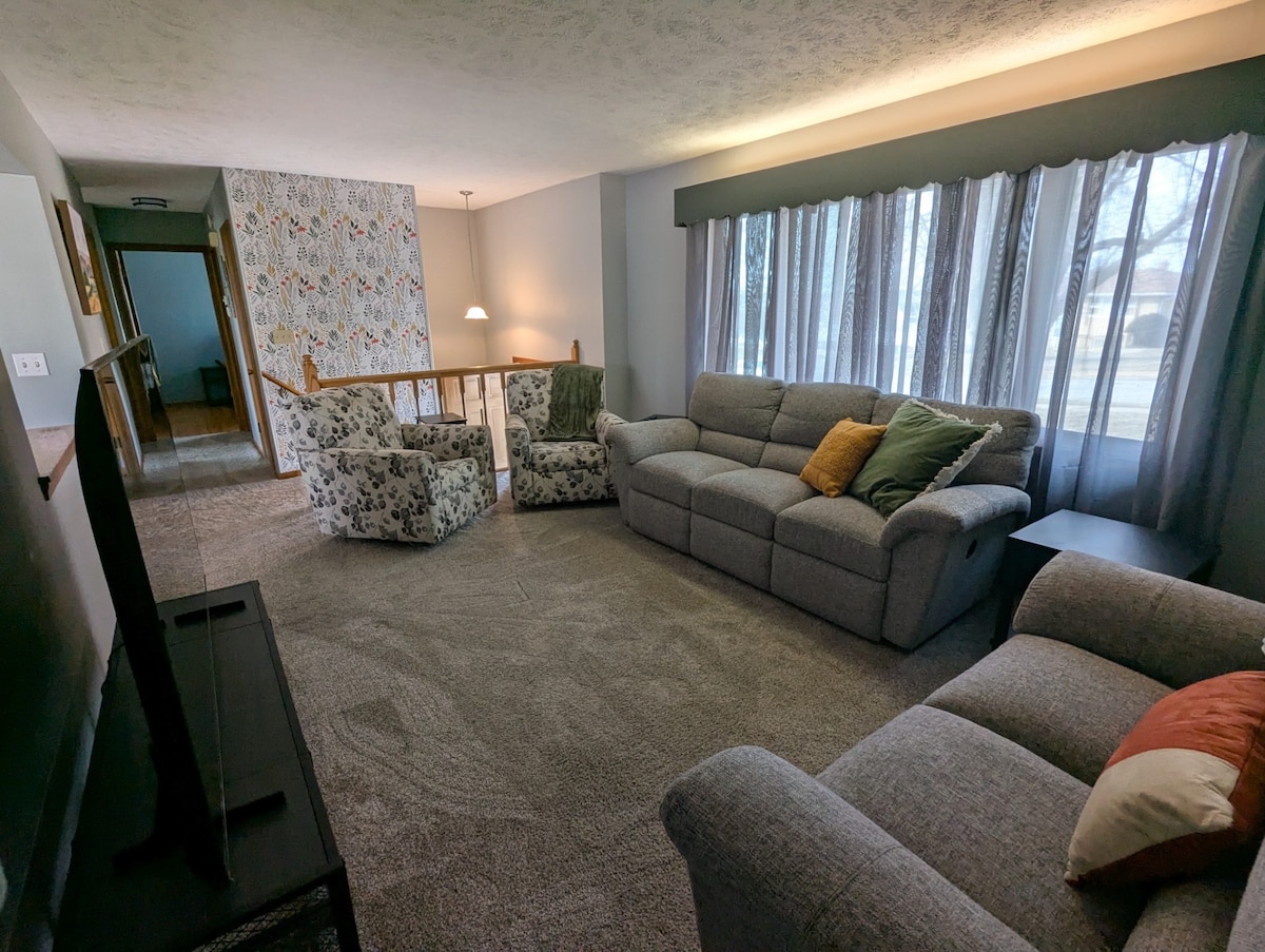 A cozy living area features soft carpeting and comfortable seating, including a spacious gray sofa and two patterned armchairs. Natural light fills the room through a large window, accentuated by sheer curtains. A side table stands next to the sofa, adding functionality to the space.