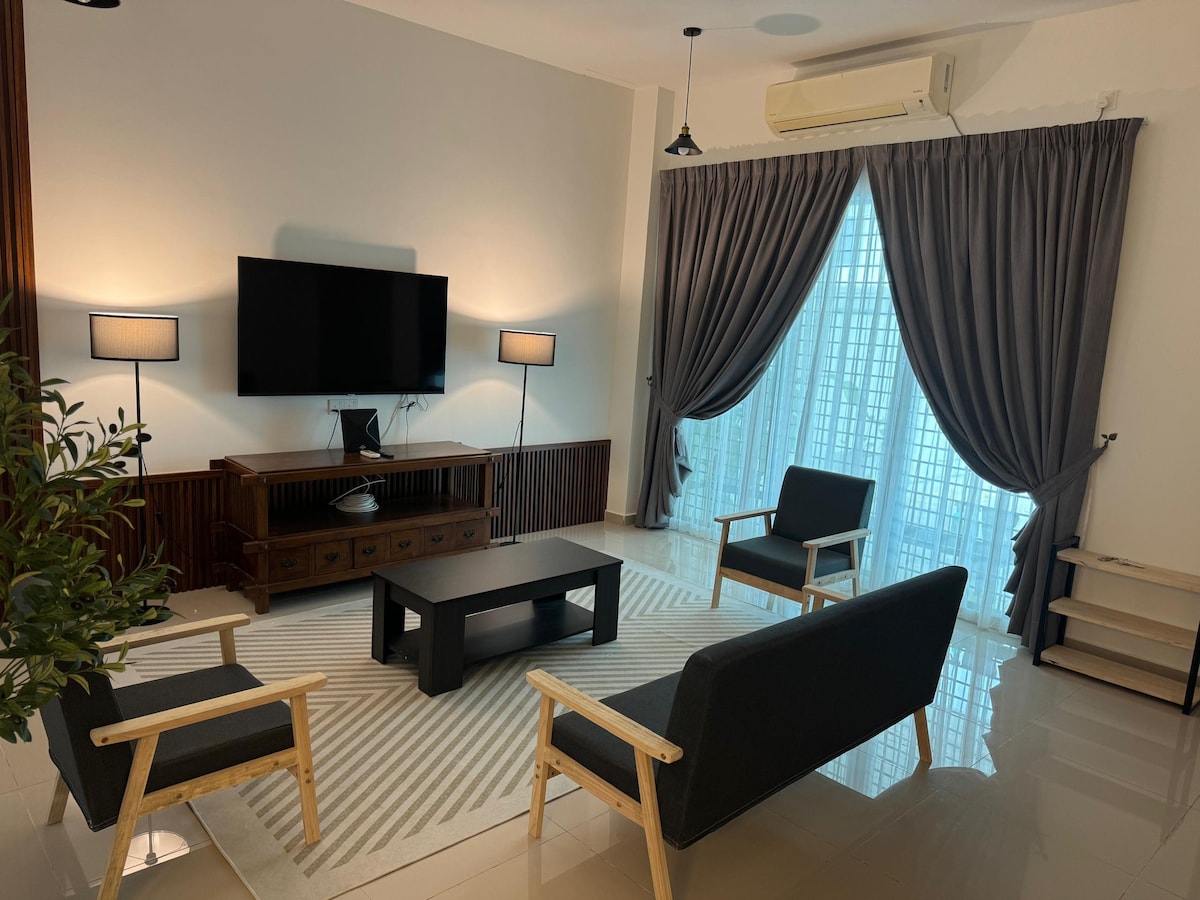 A living area is furnished with two armchairs and a sofa, arranged around a low coffee table. Natural light filters through sheer curtains, illuminating the space. A large television is mounted on the wall, complemented by decorative lamps and a potted plant.