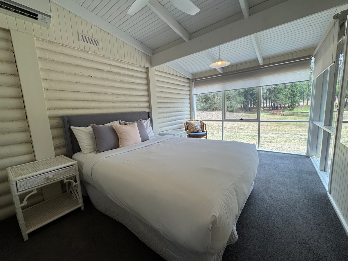 A spacious bedroom features a large bed adorned with neutral-colored pillows. An inviting chair is positioned near a window that overlooks the grassy outdoor area. Natural light filters through the sheer window treatments, enhancing the calming ambiance.