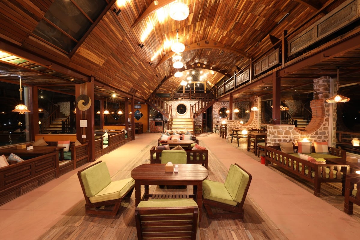 A spacious communal area features wooden beams and a high ceiling, illuminated by hanging lights. Several seating arrangements are visible, including low wooden tables surrounded by comfortable chairs, creating an inviting space for relaxation and socializing.