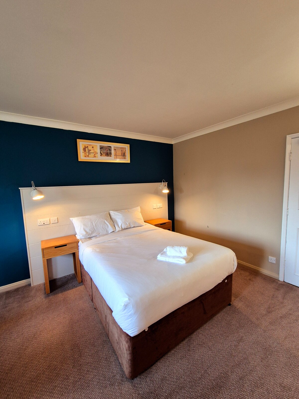 A modern sleeping area is featured, with a spacious bed dressed in white linens. Two bedside tables with lamps are positioned on either side. The walls are painted in a two-tone scheme, with one wall in deep blue and the other in a neutral shade.