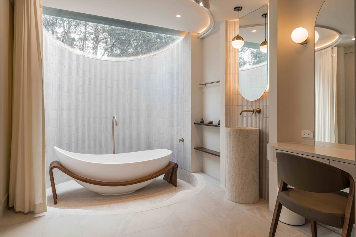A spacious bathroom features a modern freestanding bathtub situated under a circular window, allowing natural light to illuminate the space. A stone countertop with a basin and a mirror is complemented by minimalistic shelving. Soft, neutral tones create a calming environment.