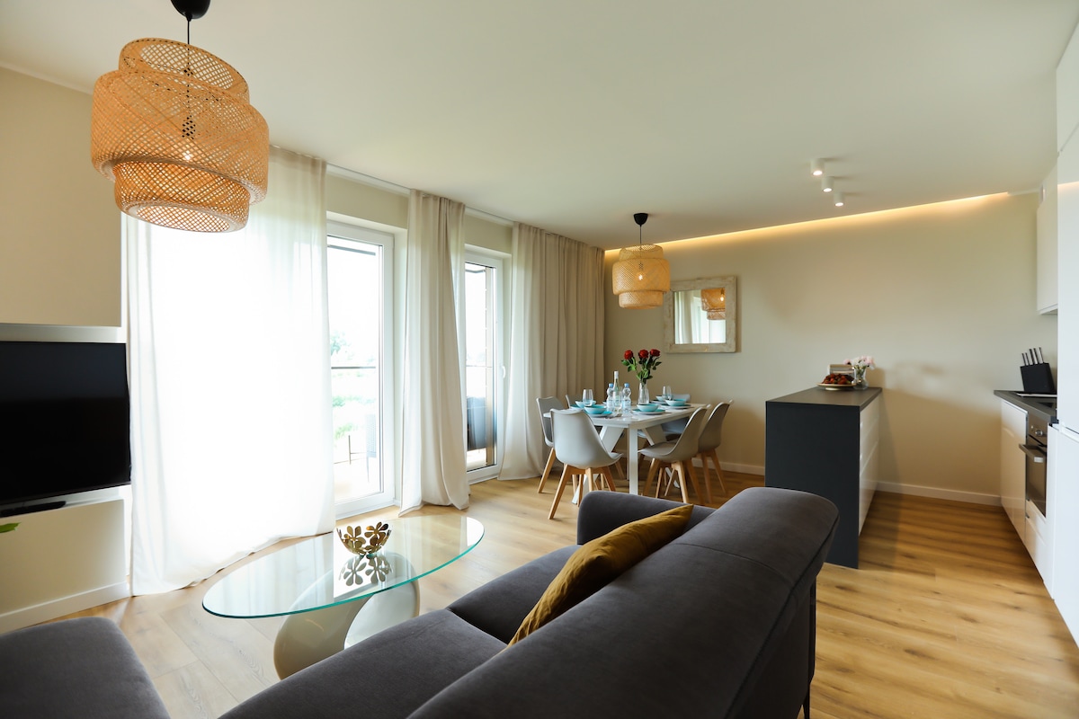 A modern living space is shown, featuring a grey sofa and a glass coffee table. Natural light enters through two large windows, flanked by sheer curtains. A dining area with a round table is set for a meal, accompanied by light wood chairs and decorative elements.