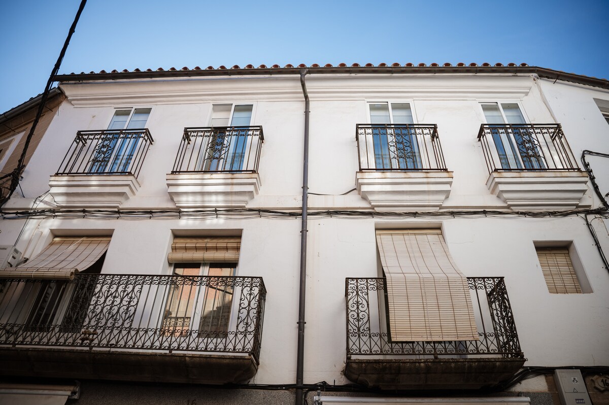 The building's façade features a classic design with a white exterior and decorative wrought-iron balconies. Large windows allow for natural light, and some are fitted with awnings for shade. The structured lines and architectural details reflect the charm of the surrounding area.