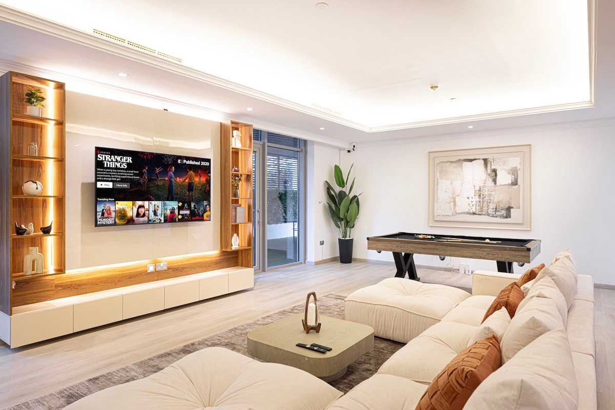 A spacious living room is furnished with a large, comfortable sectional sofa facing a wall-mounted television displaying a popular series. A sleek coffee table is centered on the light-colored flooring, while a modern pool table occupies one corner. Bright natural light enters from large sliding doors.