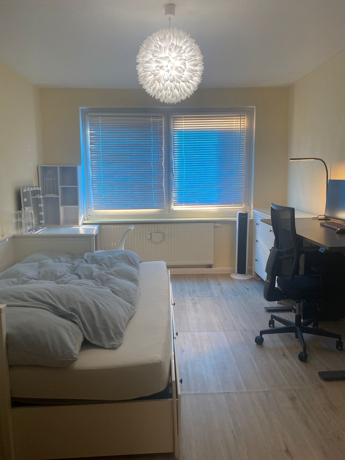 A well-lit spare room features a spacious bed with light grey bedding and a height-adjustable desk with a monitor. A makeup table is positioned nearby, and a chair sits in front of the desk. Large windows allow natural light to fill the space, complemented by a modern pendant light.