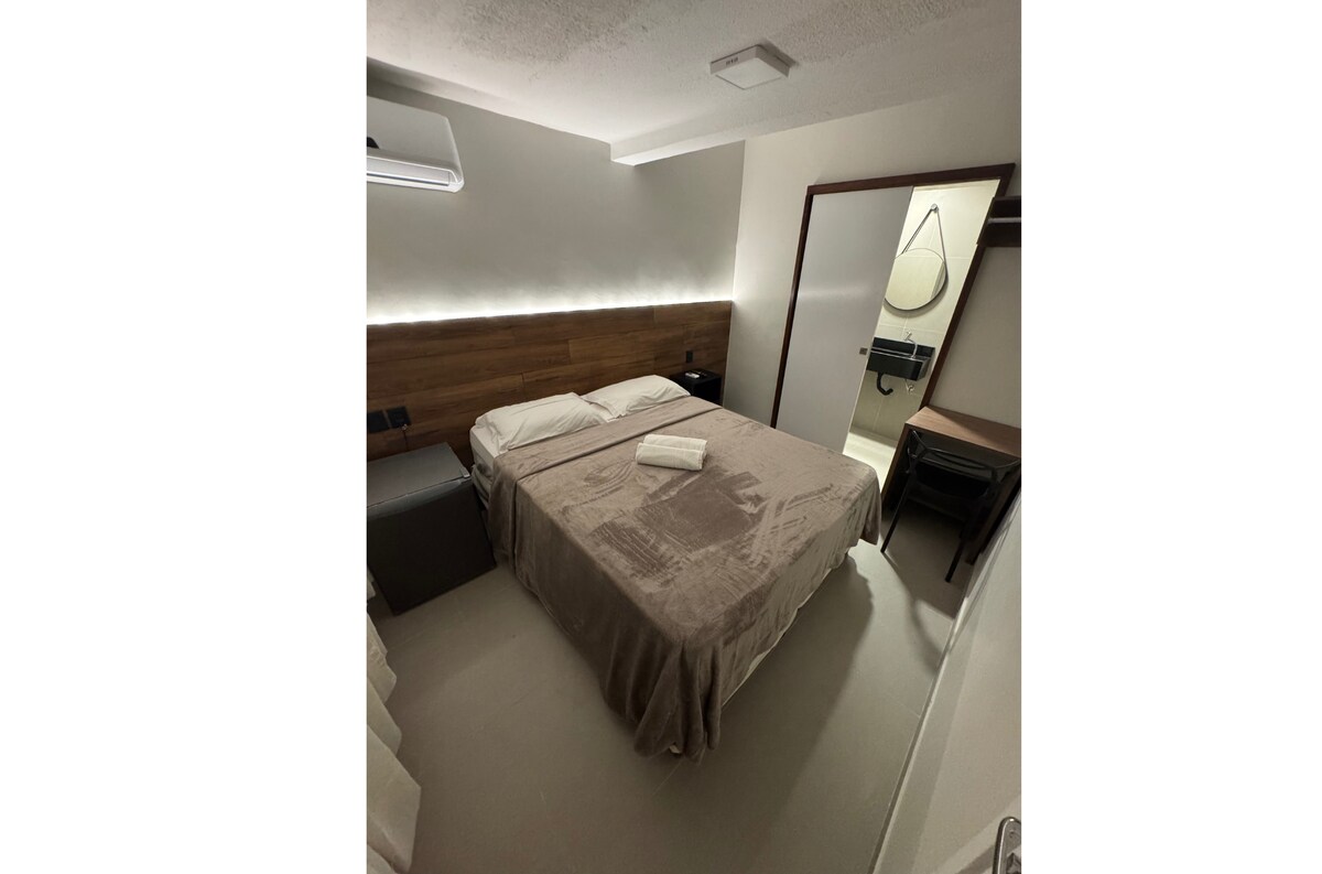 A cozy bedroom features a spacious bed with neutral bedding, situated against a wooden headboard. Subtle lighting along the wall creates a soft ambiance. A mirror and bathroom area can be seen in the background, complemented by a desk and two chairs.