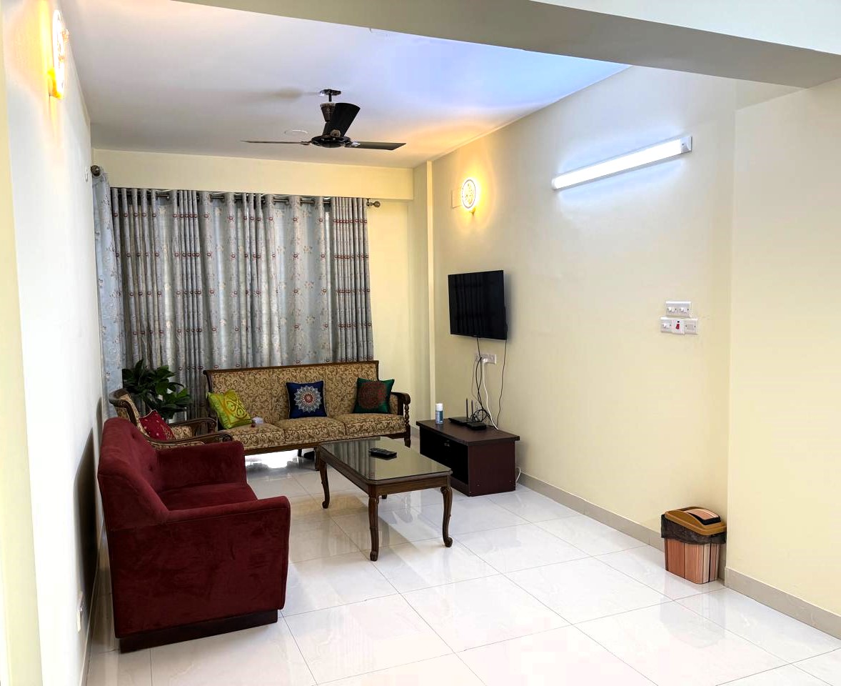 The living area features two comfortable sofa sets and a coffee table. A smart TV is mounted on the wall, and large windows with patterned curtains allow natural light. A small indoor plant adds a touch of greenery to the space.