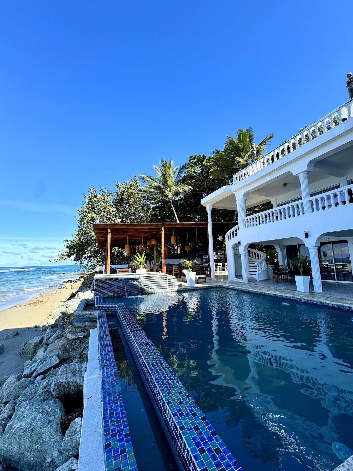 A spacious outdoor area features a clear blue pool beside the ocean. The white building with arched balconies stands against a bright blue sky and lush palm trees. A shaded sitting area is visible nearby, adding to the coastal ambiance.