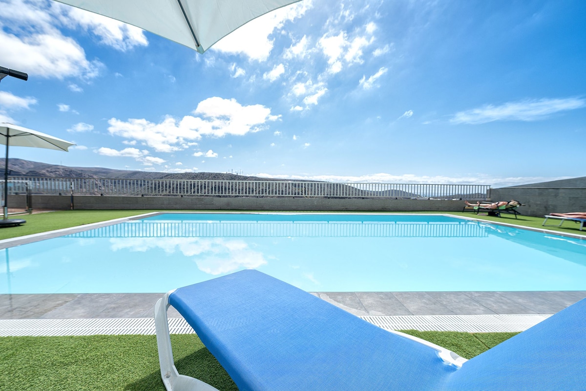 The image captures a serene swimming pool surrounded by well-maintained green grass and lounge chairs. Two umbrellas provide shade, while the clear blue sky and distant hills serve as a backdrop, creating an inviting atmosphere for relaxation.