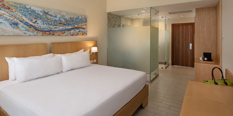 A spacious bedroom is presented, featuring a large bed with white linens and two pillows. A decorative art piece in blue tones hangs above the headboard. The room includes sleek wooden furnishings, a glass-walled bathroom area, and natural light streaming in from the entryway.