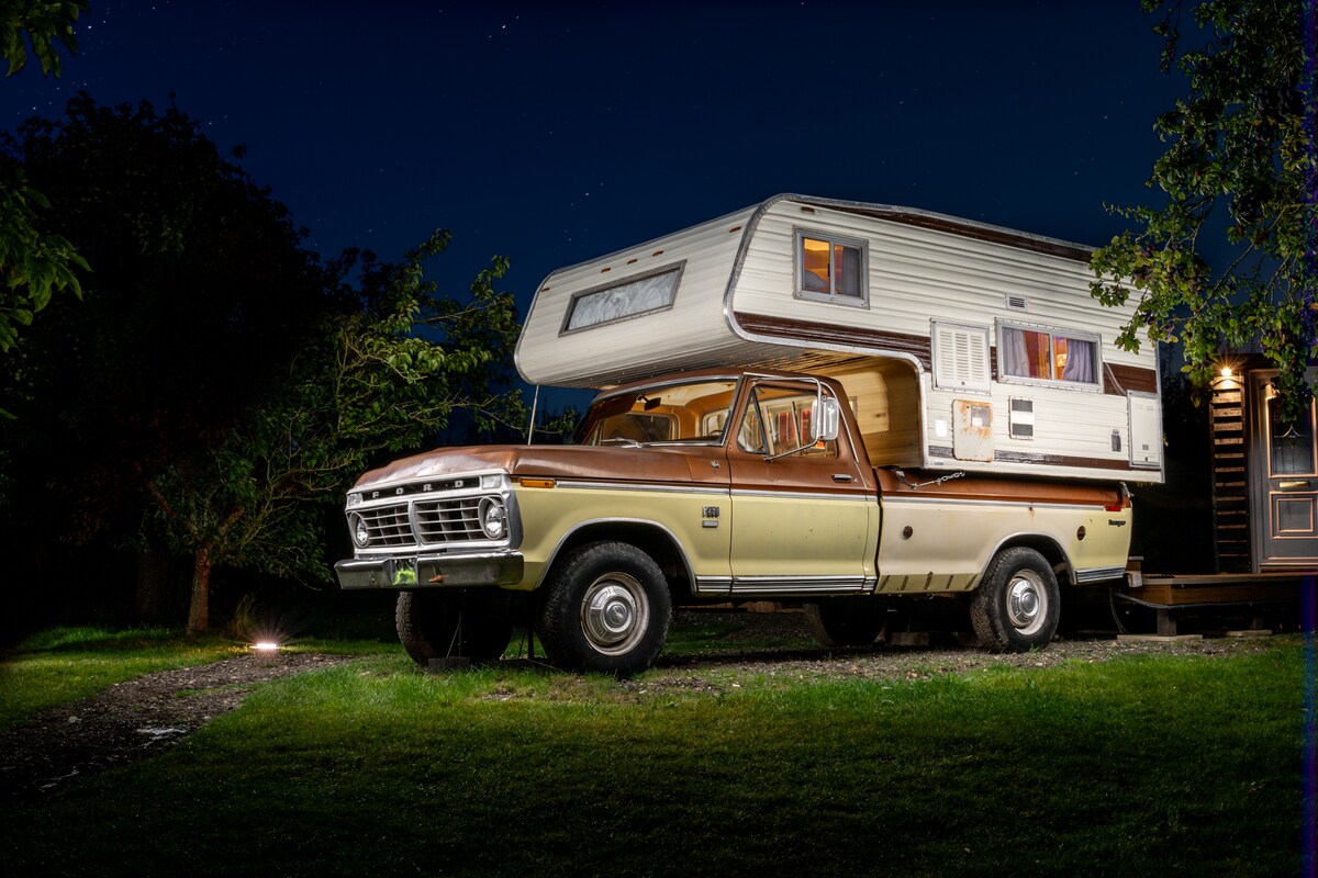 A vintage 1970s Ford F250 Camper Special is parked on a grassy area, framed by lush greenery. The vehicle features a brown and beige color scheme, with a camper unit mounted above the truck bed that includes windows for natural light. The night sky adds a serene backdrop.