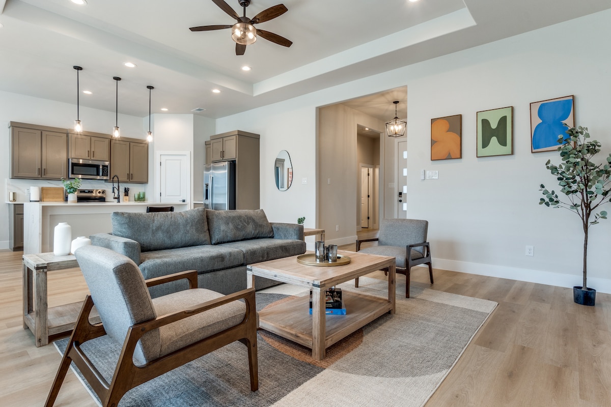 An open living area is featured with modern furnishings, including a comfortable sectional sofa and a wooden coffee table. Light-colored walls are complemented by framed art pieces. A round mirror reflects natural light, enhancing the airy space, which transitions into the kitchen area in the background.