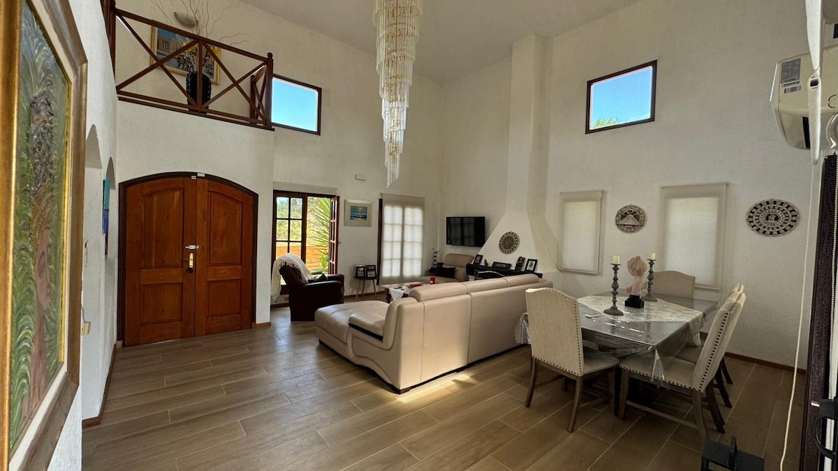 A bright living area features high ceilings and large windows, allowing natural light to fill the space. A comfortable sectional sofa is positioned near a modern media setup, while a dining table with elegant chairs is situated nearby, enhancing the open layout.