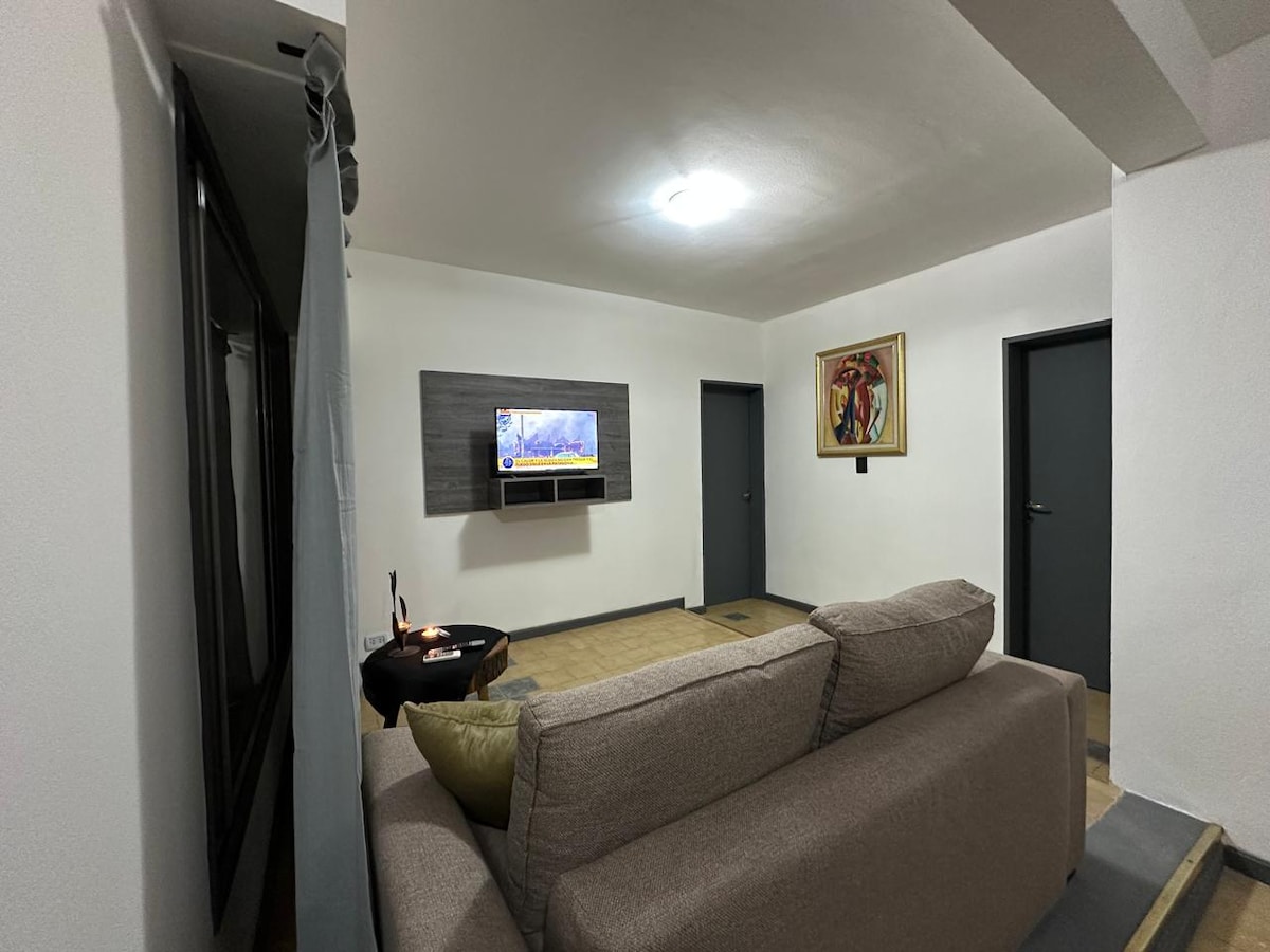 A neutral-colored living area features a comfortable sofa with accent cushions. A wall-mounted television displays a scenic image. A piece of art adorns the wall, and two closed doors are visible in the background, contributing to the room's balanced and inviting layout.