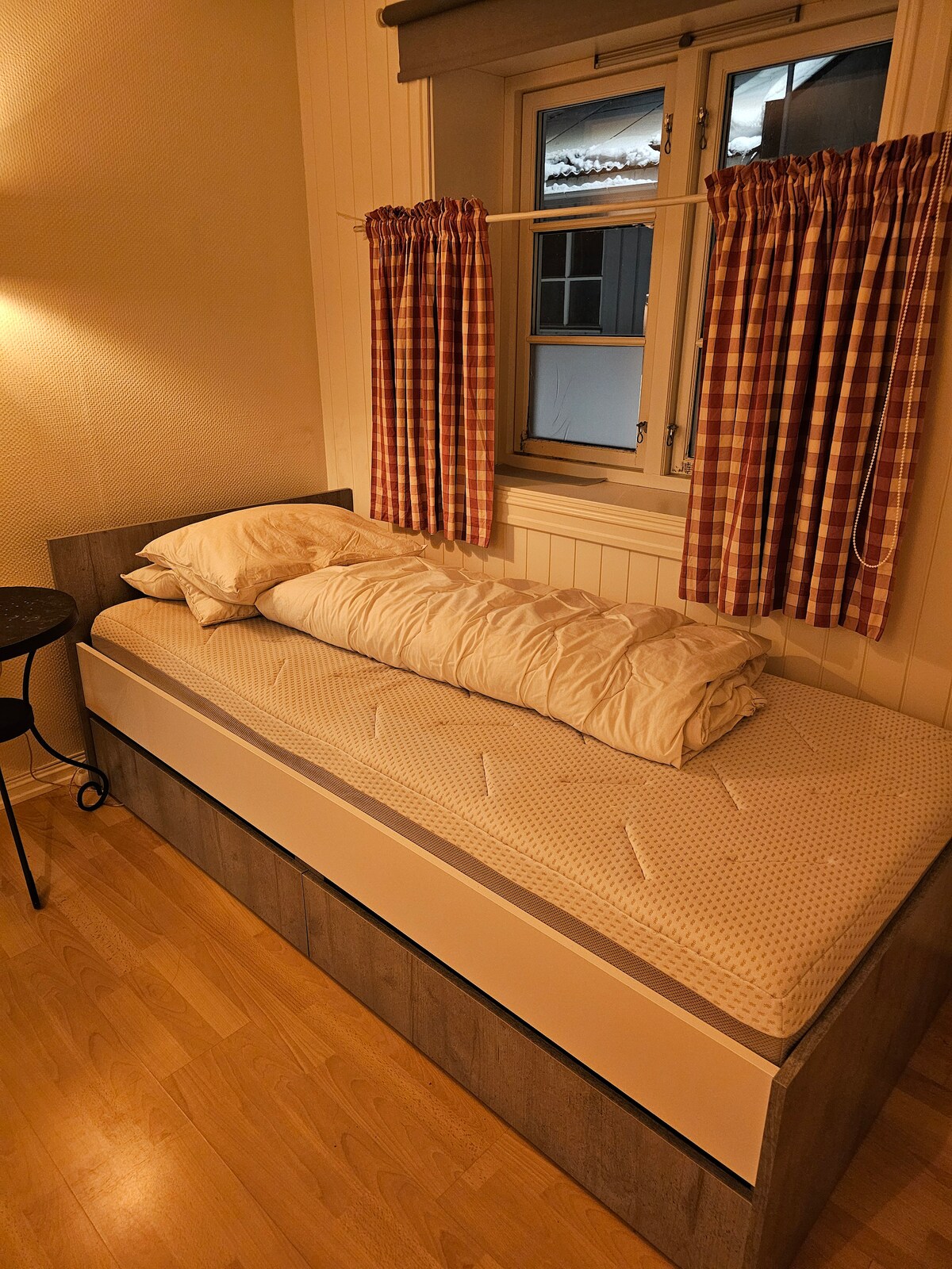A cozy sleeping area features a single bed with a light-colored mattress topped with a neatly folded blanket. Soft, checked curtains frame a window that allows for natural light, while a small round table is positioned beside the bed.