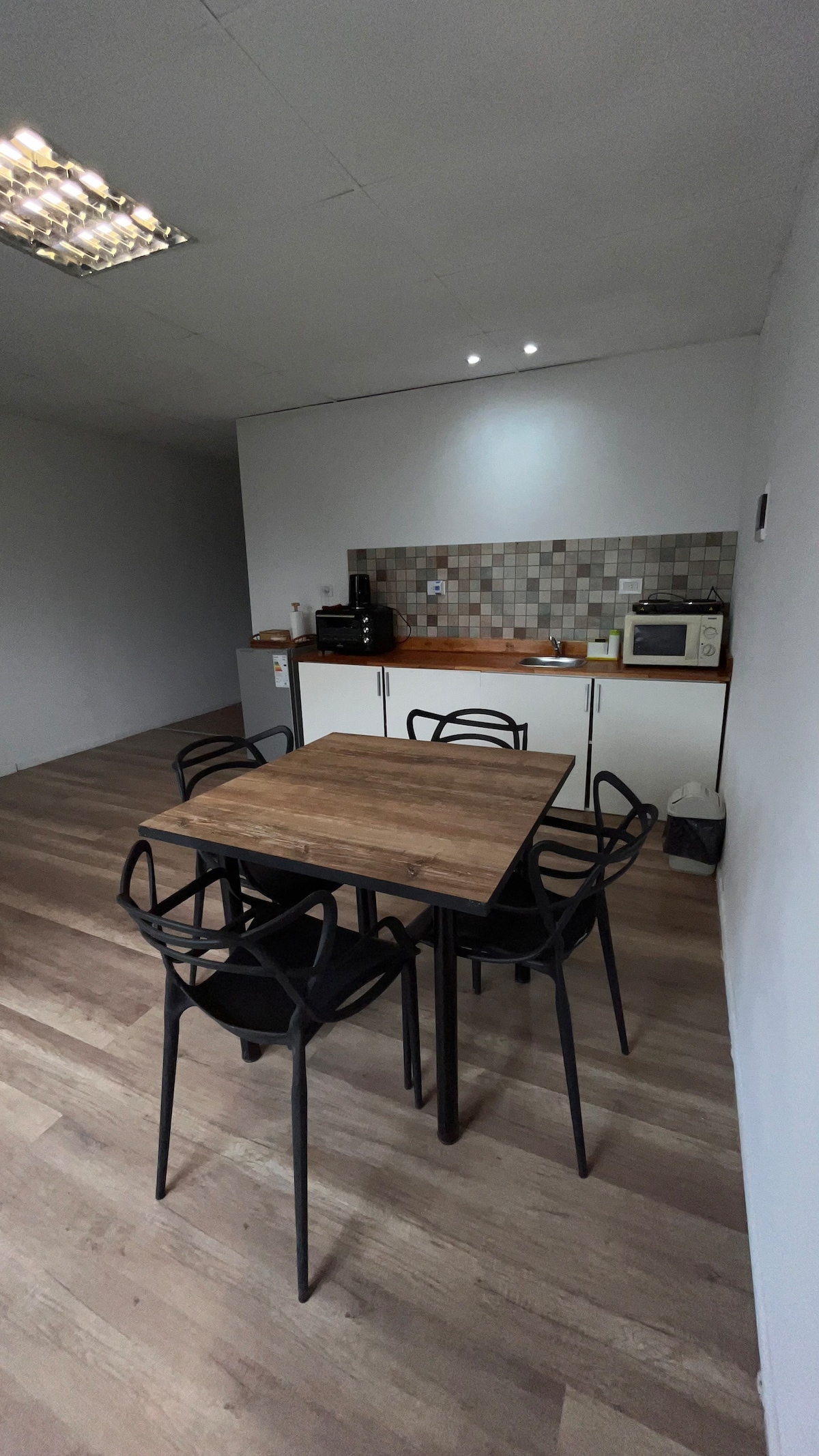 A modern kitchen space features a wooden table surrounded by four black chairs. The cabinetry is complemented by a tiled backsplash, with various appliances visible on the countertop, including a microwave and coffee maker. Natural light is softly illuminated in the area.