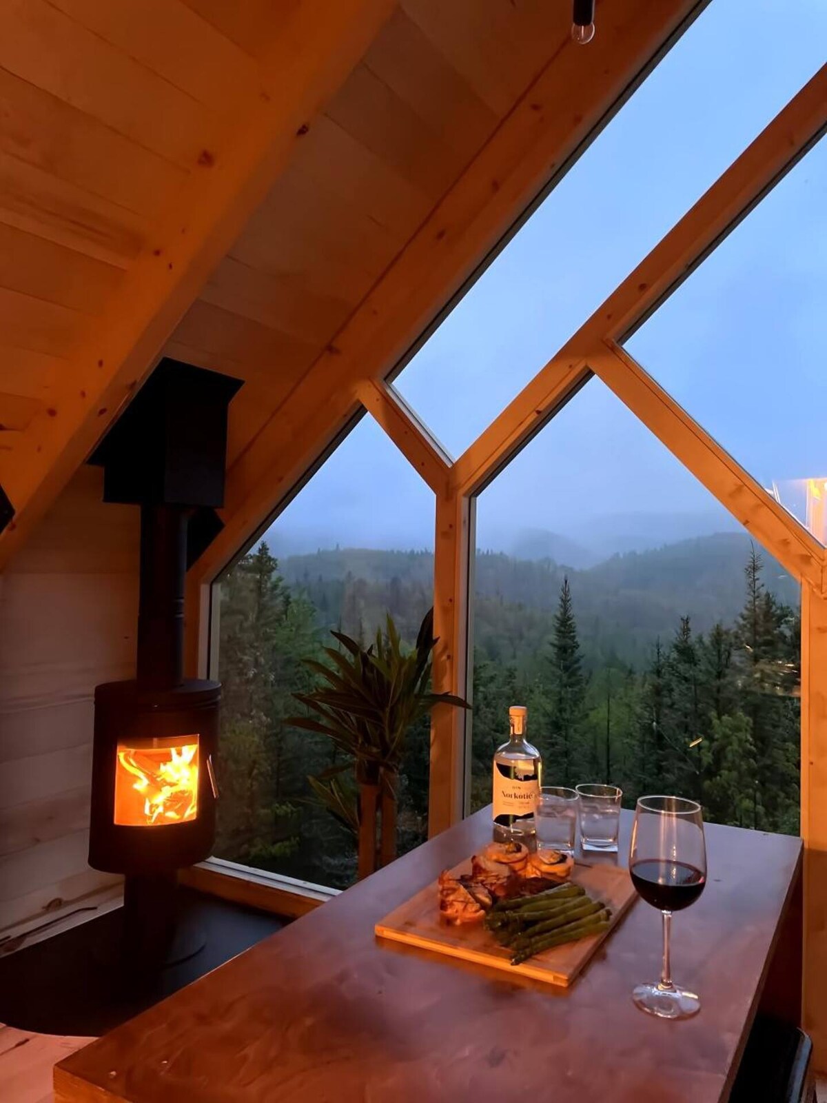 A cozy dining area features a wooden table set with a cutting board of food and drinks. A wood stove provides warmth beside large triangular windows, offering views of a misty forest landscape. Soft light casts a warm glow against the wood interior.