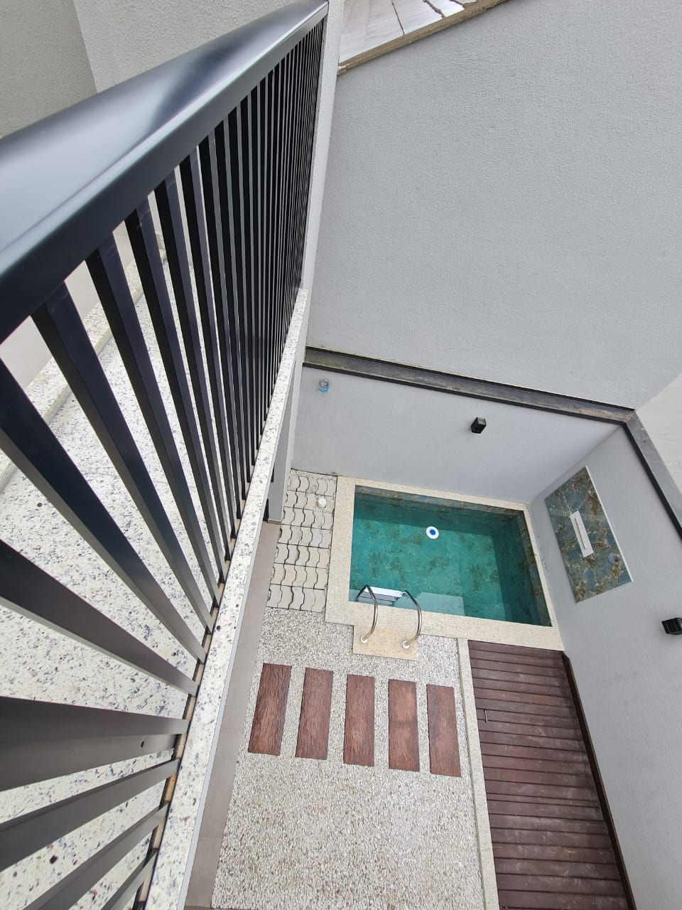 An outdoor area is visible from a balcony view, showcasing a compact swimming pool with turquoise water. Surrounding the pool, a combination of textured stone and wooden decking creates a welcoming atmosphere, while a small shower is positioned nearby.