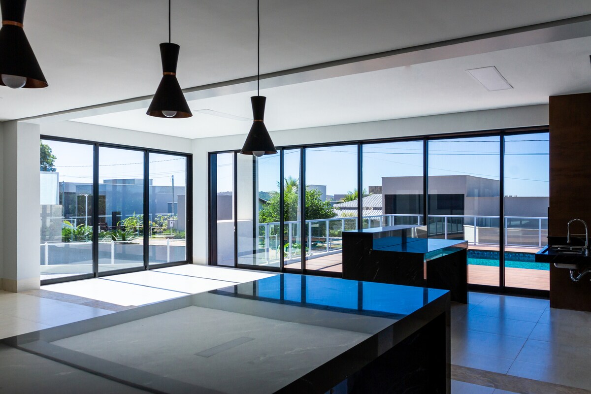 The spacious kitchen area is highlighted by large glass windows, allowing natural light to fill the room. A sleek countertop is positioned centrally, with modern pendant lights hanging above. The view of the outdoor space and pool can be observed through the expansive glass doors.