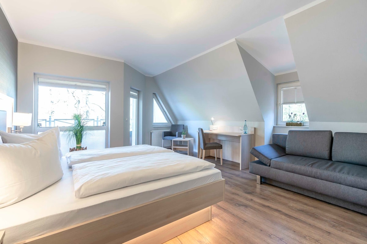 A spacious room featuring a comfortable bed and an inviting seating area with a gray sofa. Large windows allow natural light to fill the space, and a balcony is accessible from the room. A desk is also present, providing a functional work area.