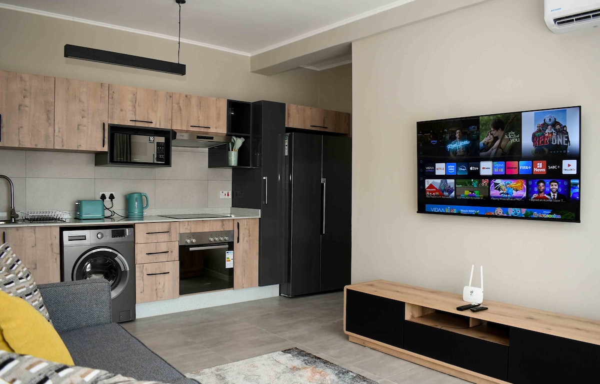 A modern kitchen area includes cabinetry in warm wood tones and sleek black accents, with a stainless-steel refrigerator and built-in appliances. A comfortable seating area is adjacent, featuring a sofa with decorative pillows. A wall-mounted television displays various streaming options.