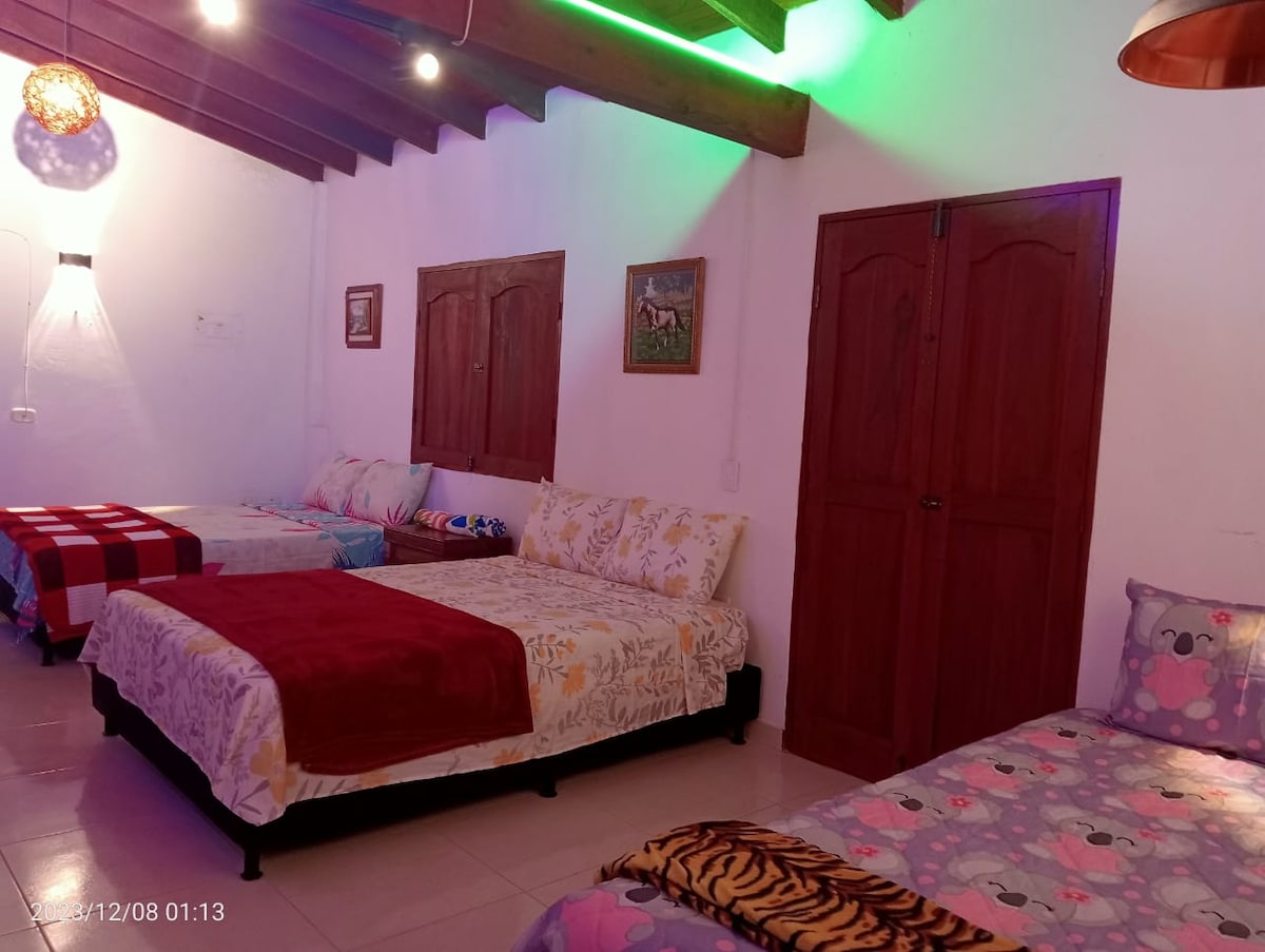 A cozy sleeping area is arranged with multiple spacious beds, each adorned with colorful blankets and cushions. The room features warm wooden beams along the ceiling, and soft lighting adds a gentle glow to the white walls. Artwork is displayed on the walls.