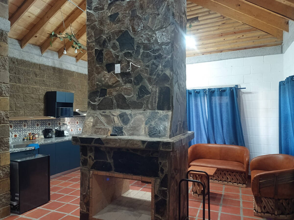 A stone fireplace serves as the focal point in a cozy living area, surrounded by wooden beams and terracotta tiles. A small kitchenette featuring dark cabinetry is visible, while two brown leather sofas are positioned nearby, complemented by blue curtains framing the window.
